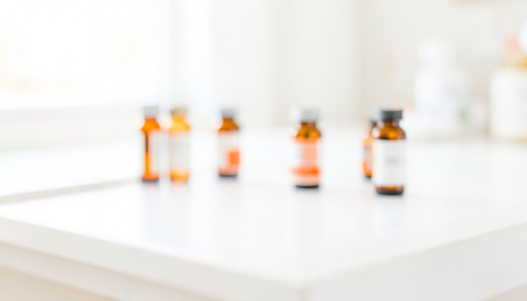 A bright, airy photograph of a clean white pharmacy countertop with a few small amber prescription bottles softly blurred in the background